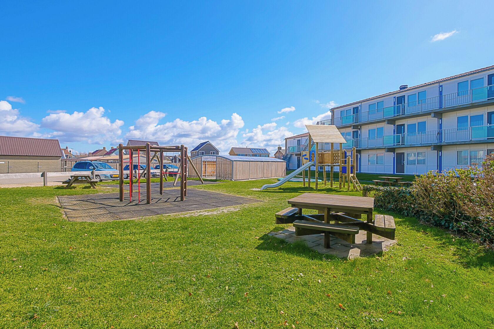 Ein Spielplatz mit Rutsche und Schaukeln, Picknicktischen und einem Gebäude mit Balkonen im Hintergrund.
