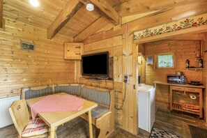 A wooden cabin interior with a dining table, bench seating, a television, a small kitchenette, and a decorative wall panel.