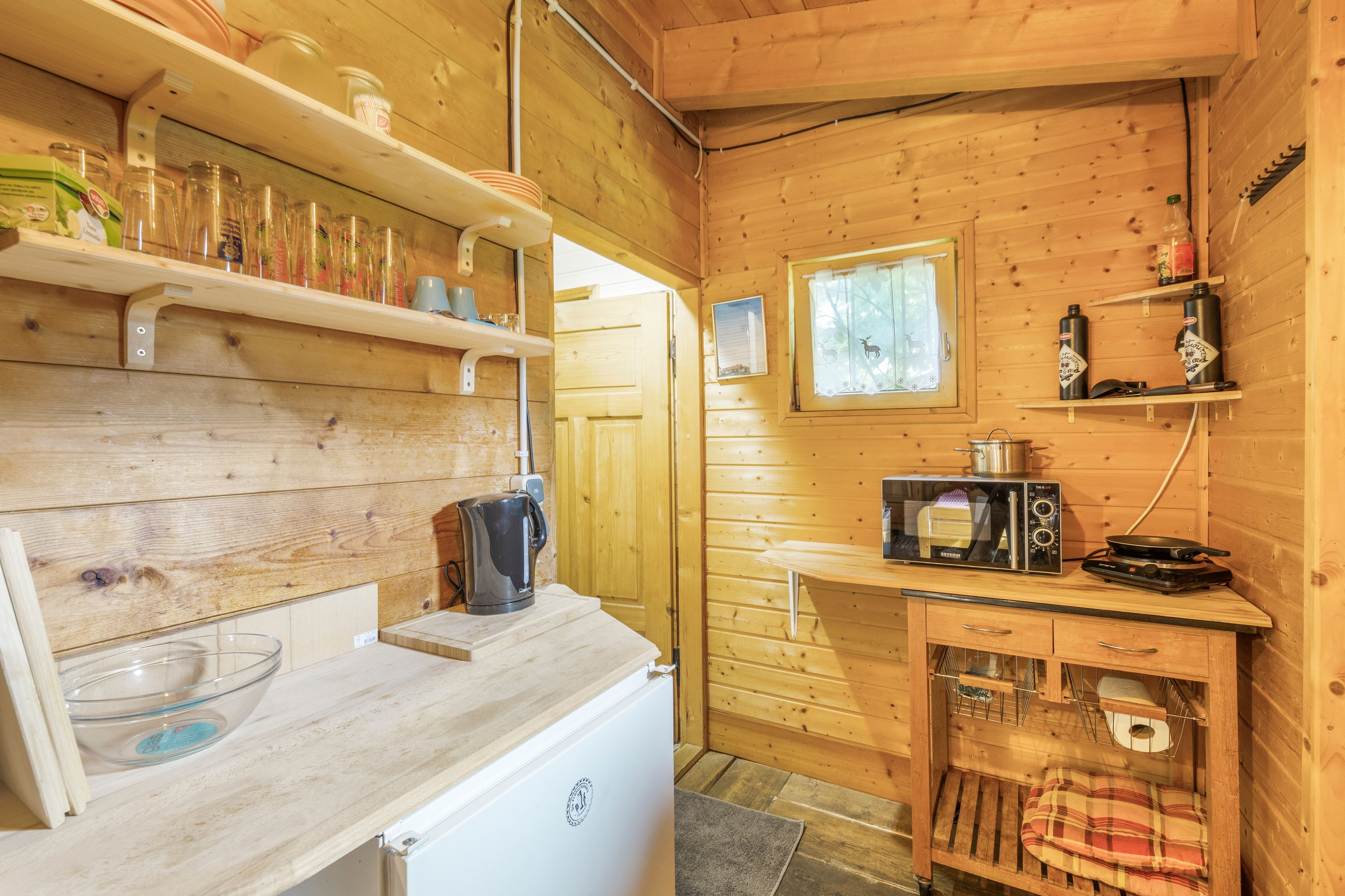 Eine kleine Küche mit Holzwänden, einer Mikrowelle, einem Wasserkocher und verschiedenen Küchenutensilien in Regalen.