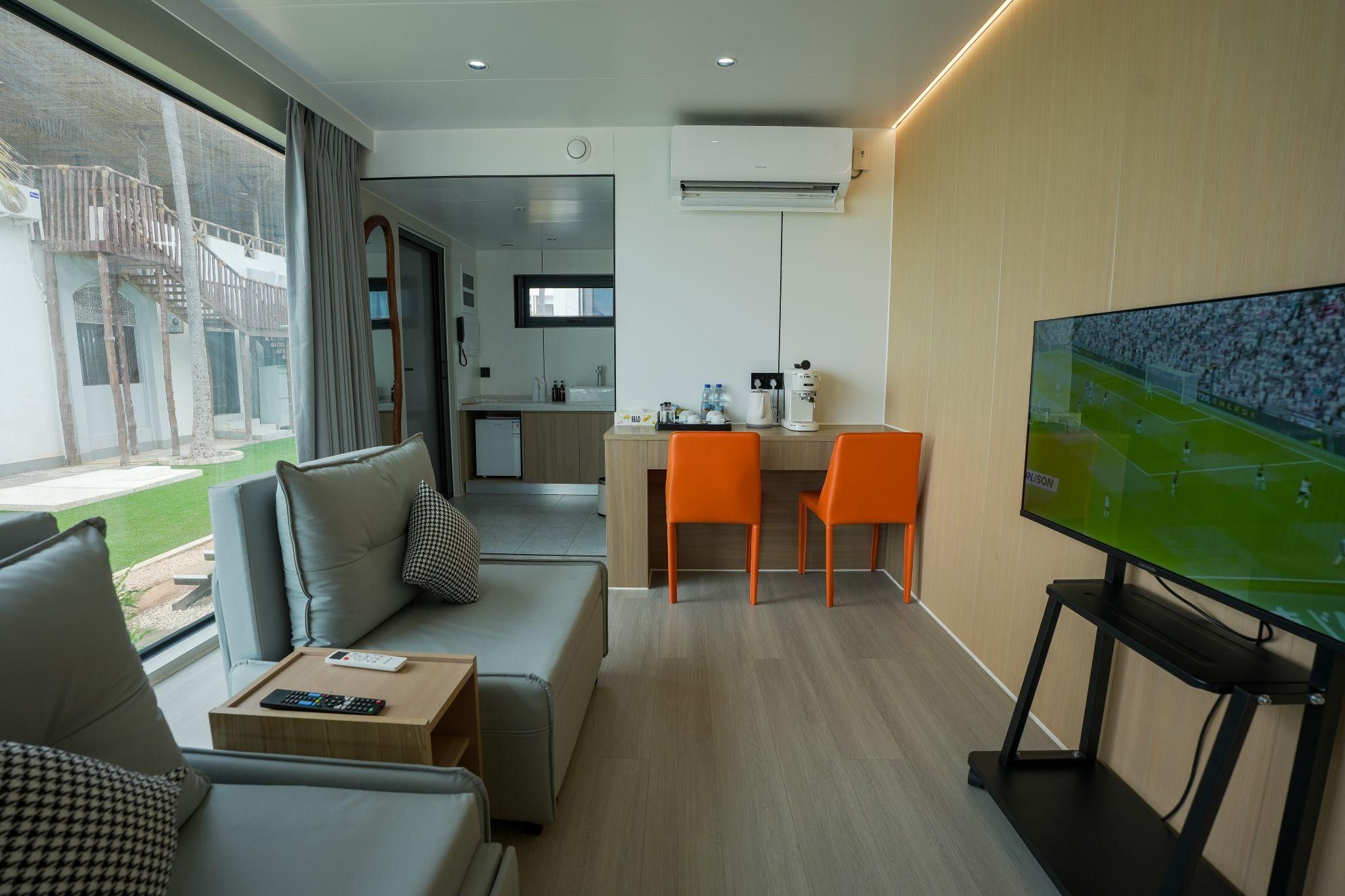 Una habitación de hotel moderna con sofá, televisión, escritorio con sillas, área de cocina y balcón con vistas.