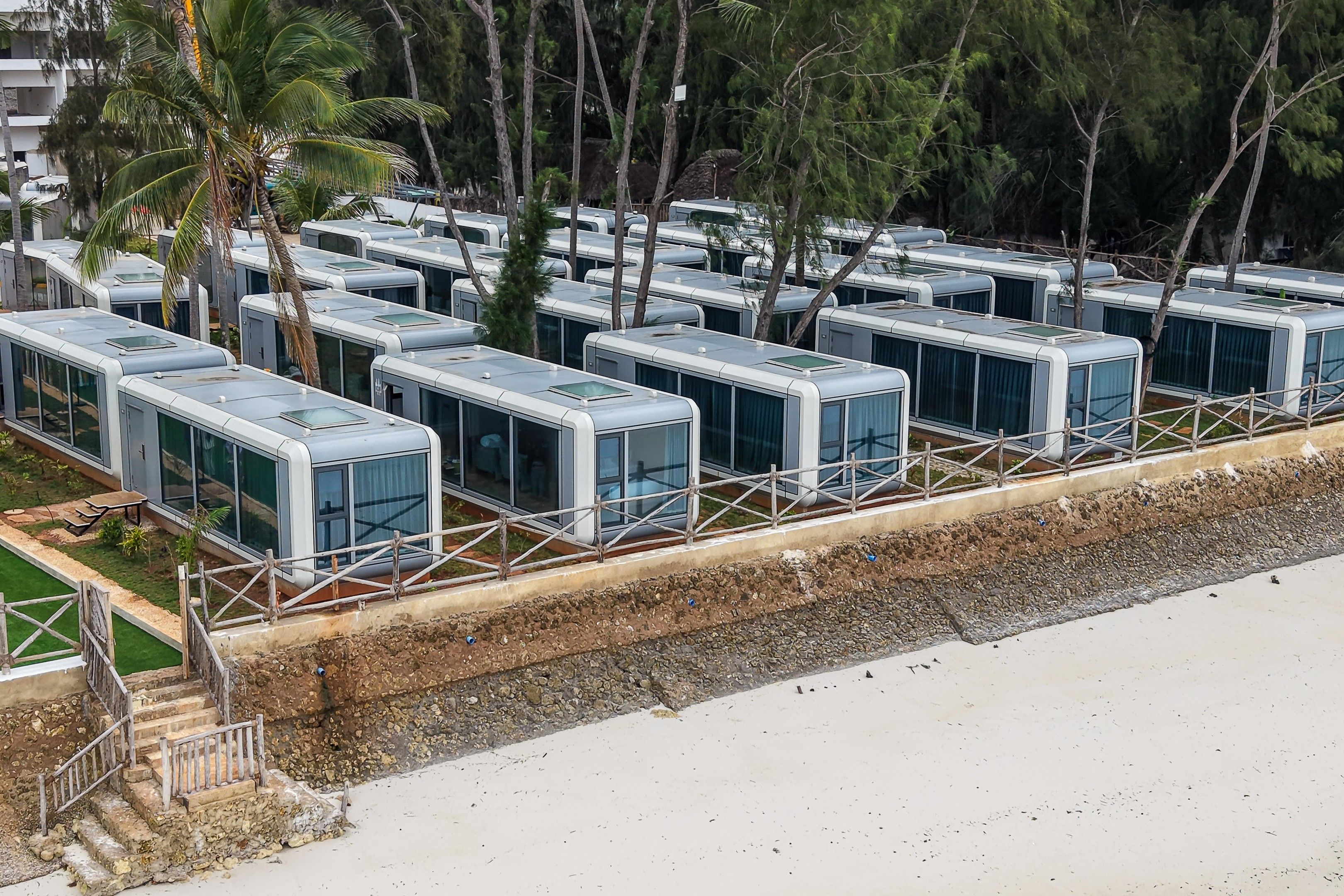 Une rangée de cabines modernes et compactes, aux murs de verre et au toit plat, est aménagée le long d’une plage, avec une digue rocheuse et une végétation clairsemée.