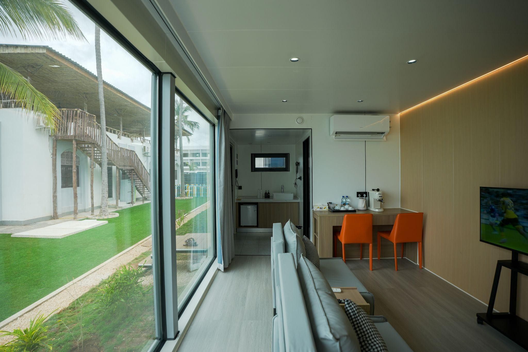 Une chambre d’hôtel moderne dotée d’une grande fenêtre, d’un coin salon avec des fauteuils orange, d’un téléviseur à écran plat et d’une kitchenette.