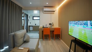 Habitación de hotel moderna con sofá, escritorio y dos sillas, televisor y una kitchenette al fondo.
