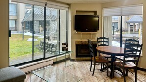 A modern living room with a fireplace, a dining table with chairs, a flat-screen TV mounted on the wall, and large windows offering a view of the outdoors. - Marquise Magic Mountain Escape. (Whistler)