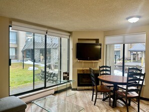 A modern living room with a fireplace, a dining table with chairs, a flat-screen TV mounted on the wall, and large windows offering a view of the outdoors. - Marquise Magic Mountain Escape. (Whistler)