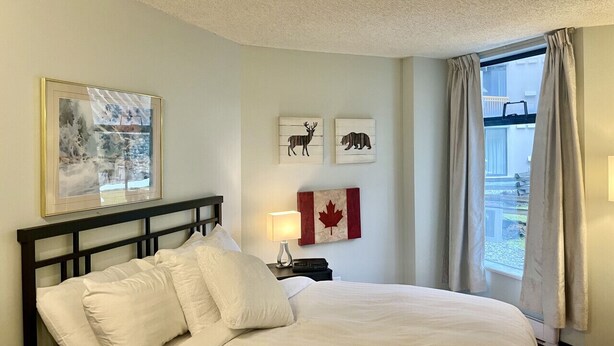 A hotel room with a bed, a nightstand with a lamp, a framed picture, and a window with curtains. - Marquise Magic Mountain Escape. (Whistler)