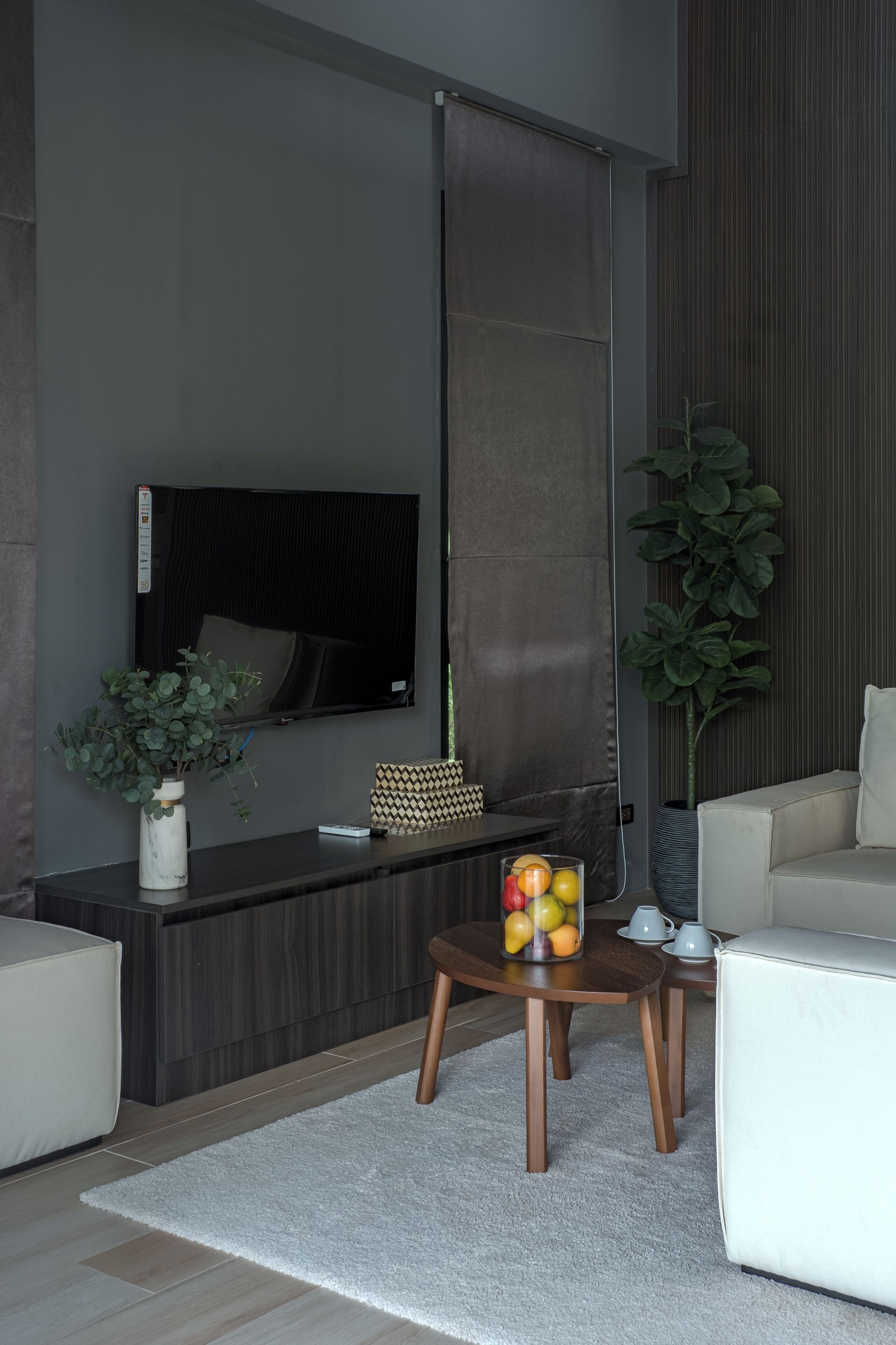 Un salon moderne avec un téléviseur à écran plat fixé au mur, une petite table basse en bois sur laquelle sont disposés des fruits, une plante en pot et un canapé.