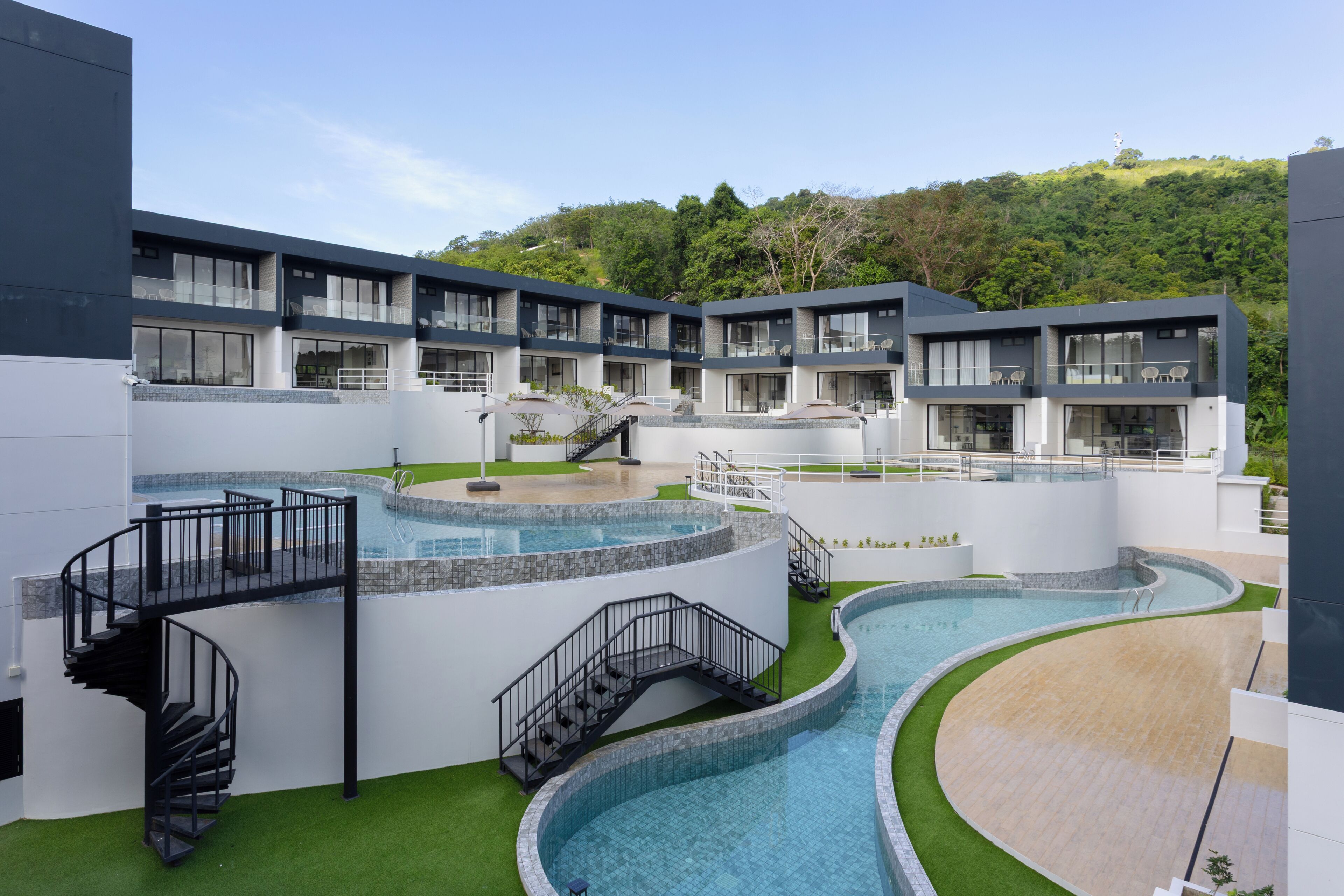 A modern hotel with multiple balconies, a central swimming pool, and a lush green hillside in the background.
