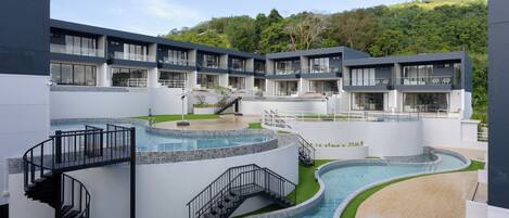 A modern hotel with multiple balconies, a central swimming pool, and a lush green hillside in the background.