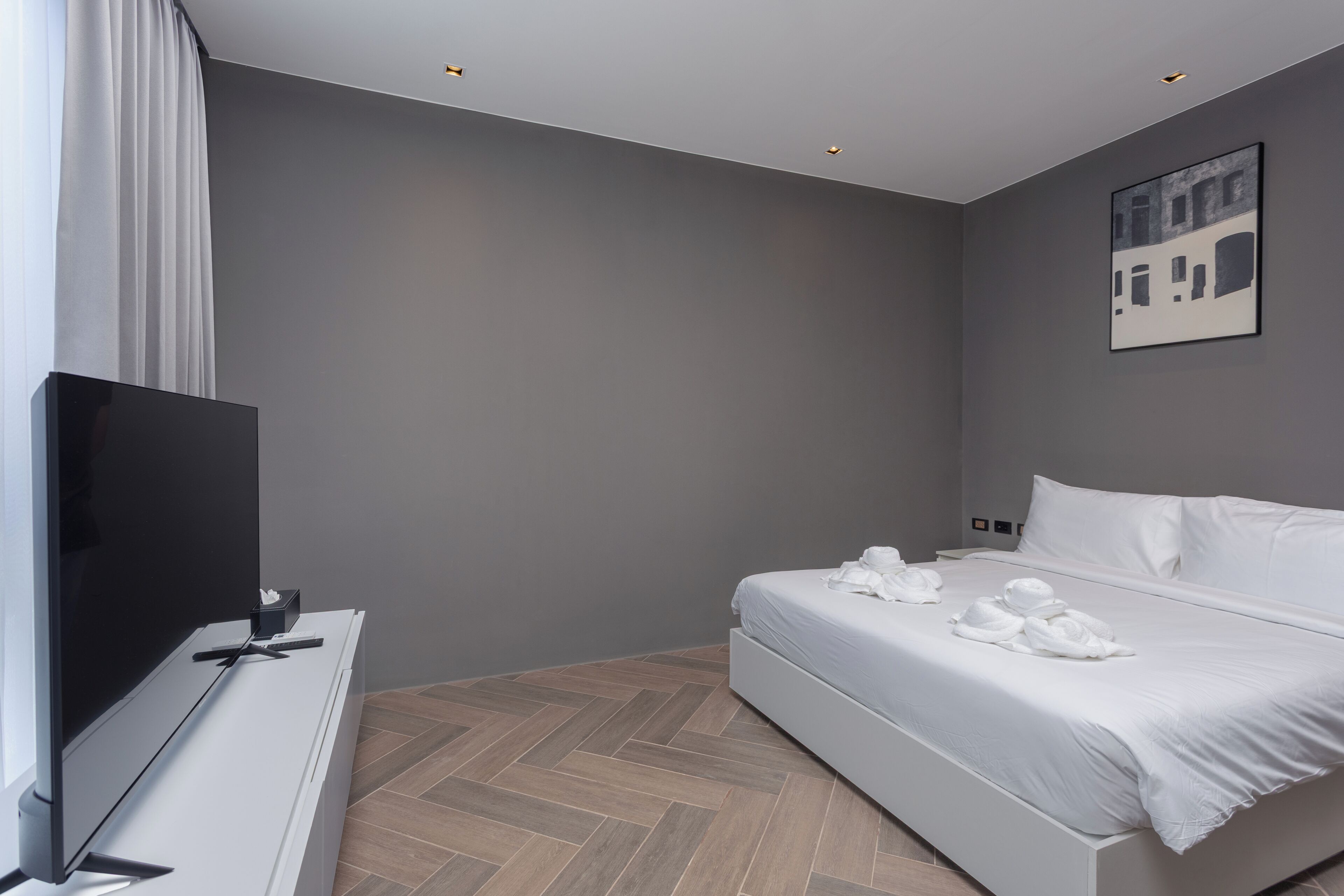 A modern hotel room with a bed, a flat-screen TV, a desk with a computer, and a herringbone-patterned floor.