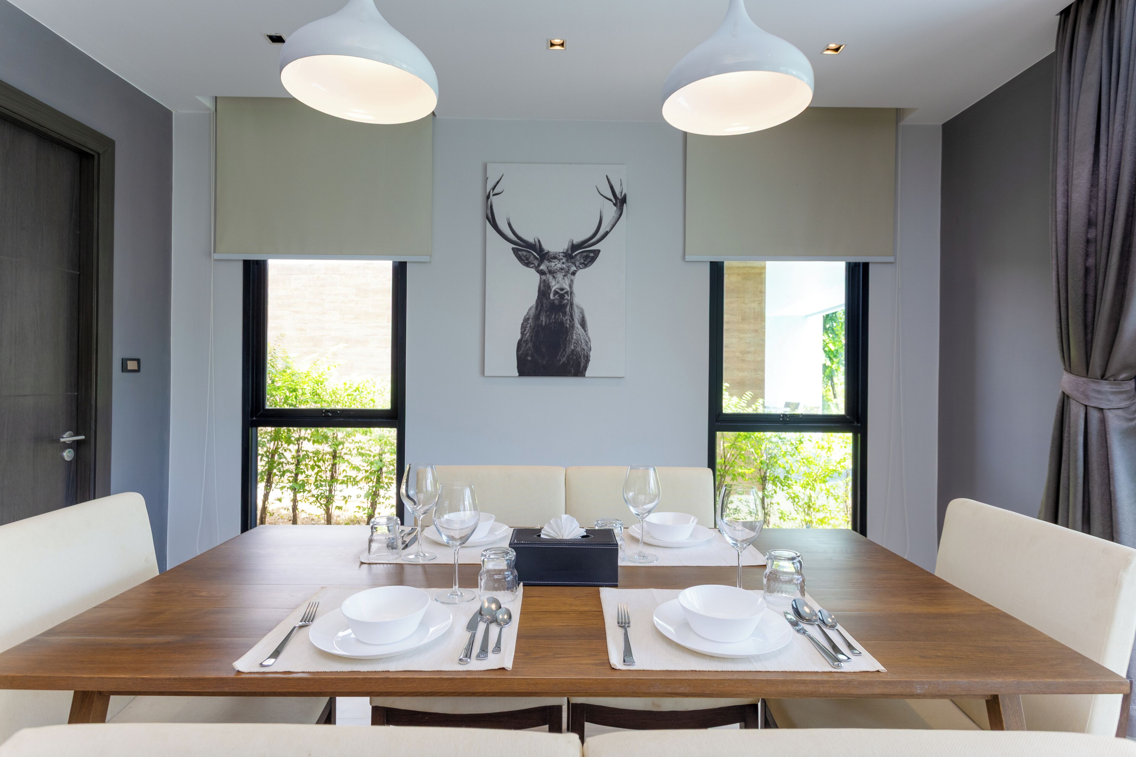 A dining room with a wooden table set for two, two pendant lights, a framed deer head artwork, and a view of greenery through the windows.