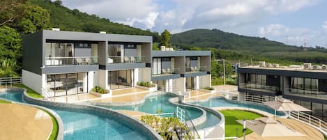 A modern resort with a large swimming pool, multiple buildings, and a mountainous backdrop.