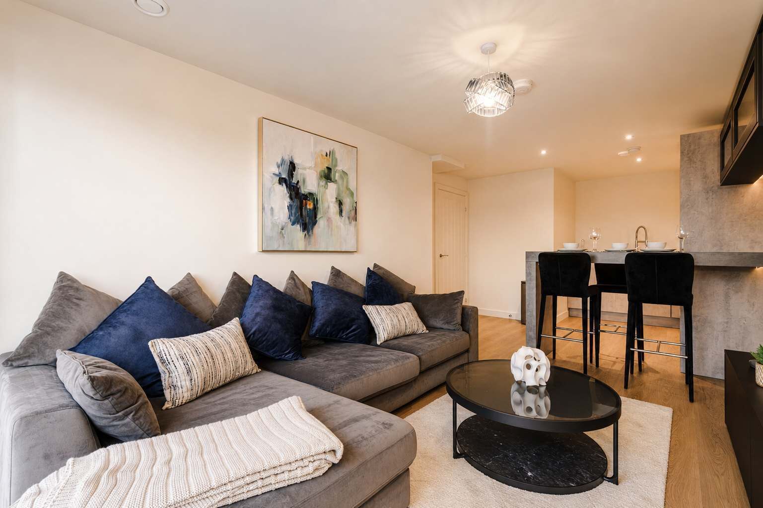 A modern living room with a grey sofa, a coffee table, and a dining area with a bar stool.
