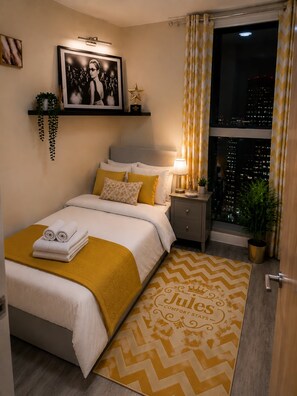 Ein Schlafzimmer mit Bett, Nachttisch, Lampe, Topfpflanze und einem Fenster mit Blick auf die Stadt.