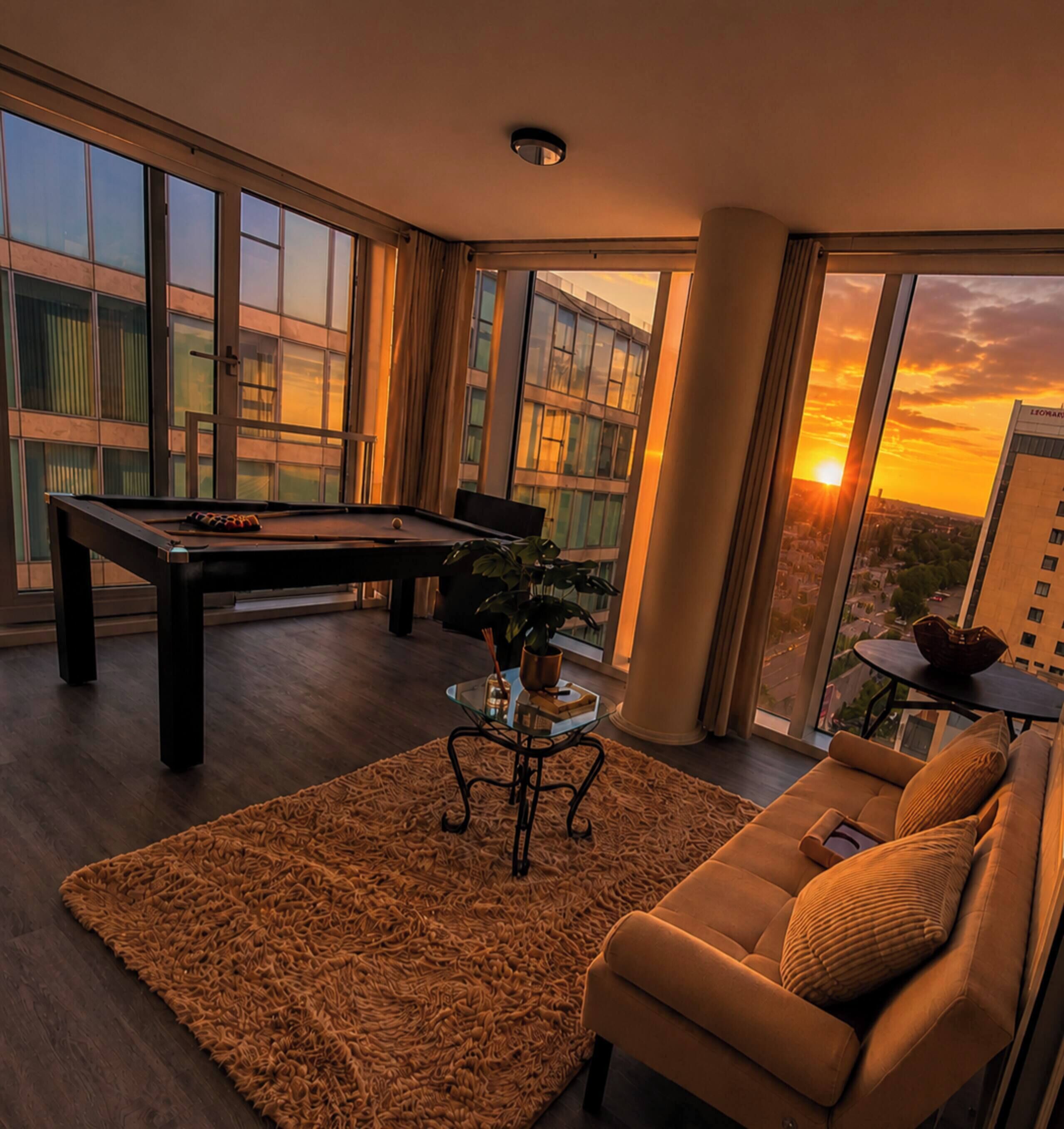 A modern living room with a pool table, a sofa, and a large window overlooking a cityscape at sunset.