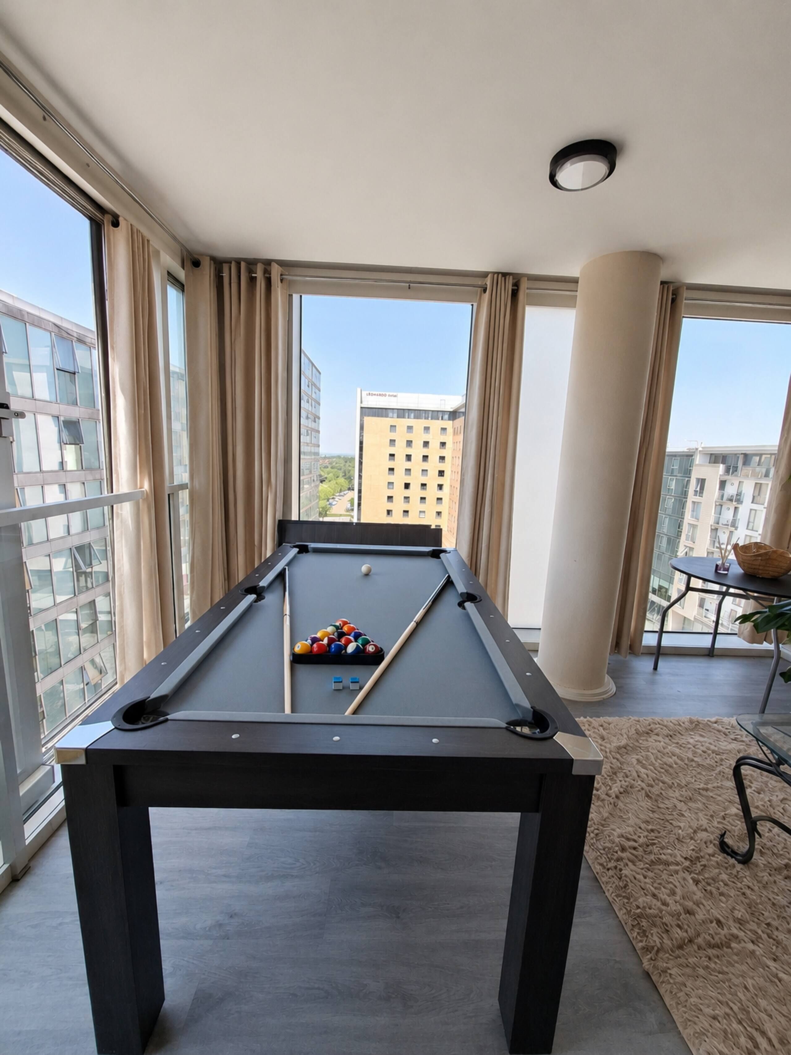 A pool table in a room with large windows, a view of buildings, and a small table with a basket.