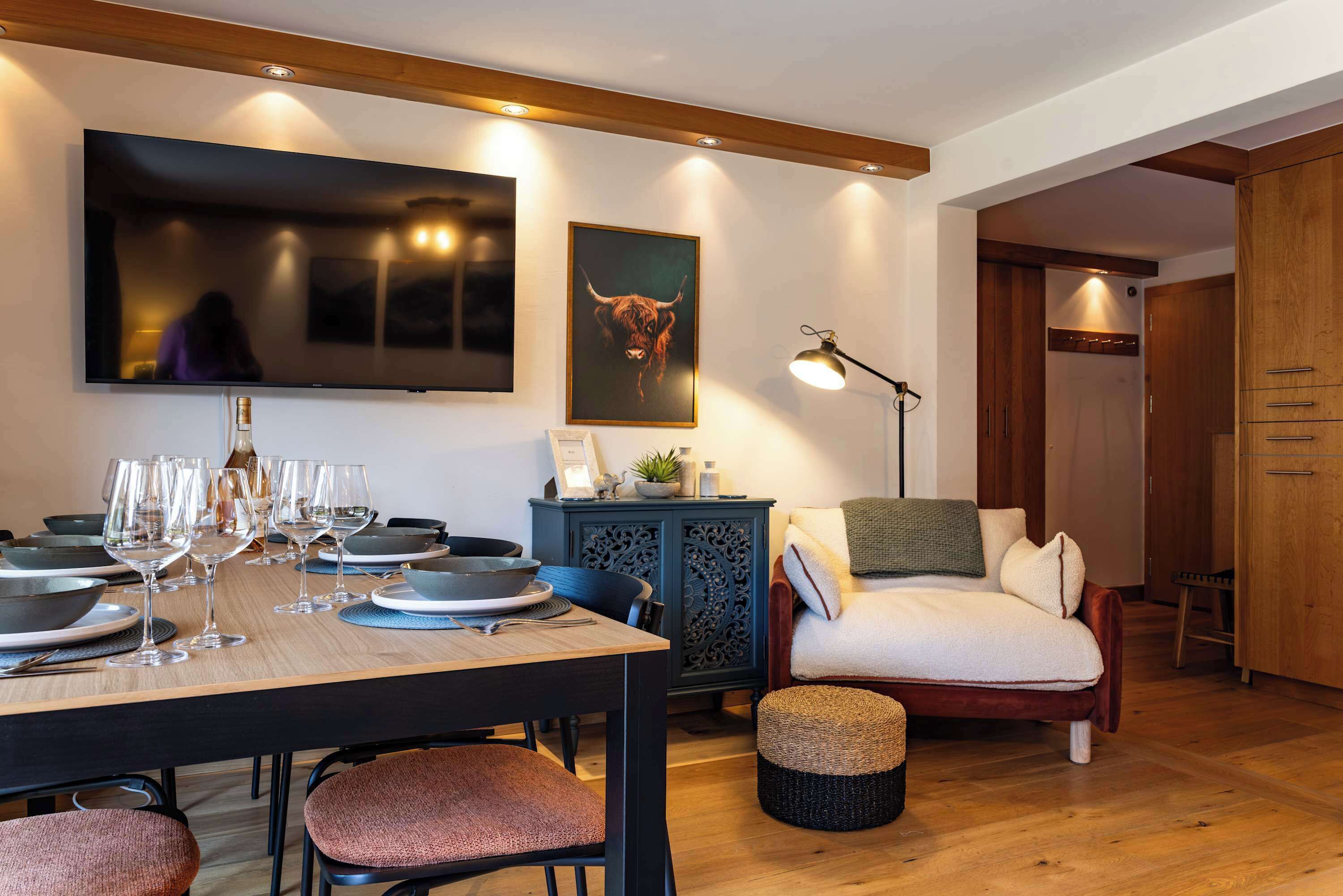 Un comedor con una mesa de madera lista para una comida, un sofá, una silla y un televisor montado en la pared.