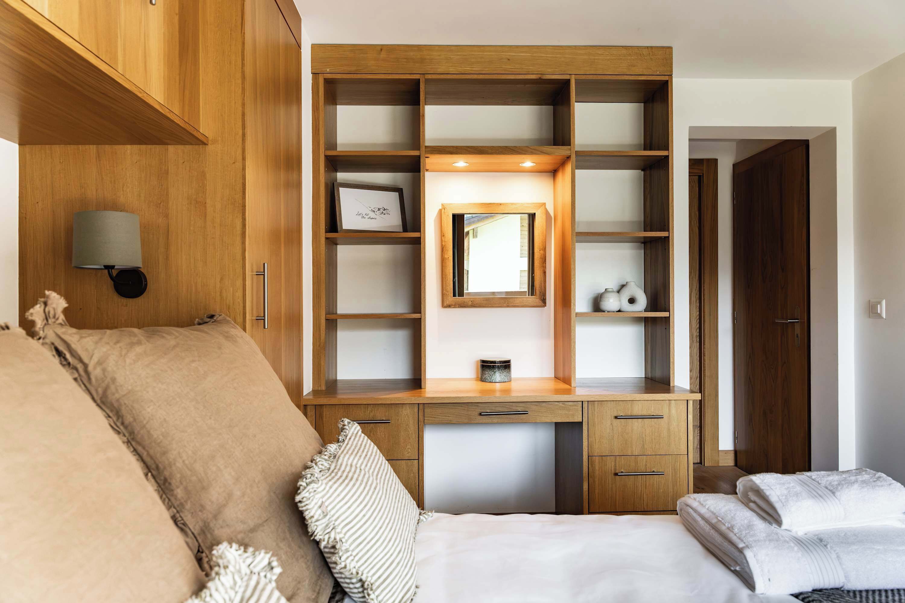 A bedroom with a wooden desk, a bed, a nightstand, a mirror, and a wall-mounted lamp.