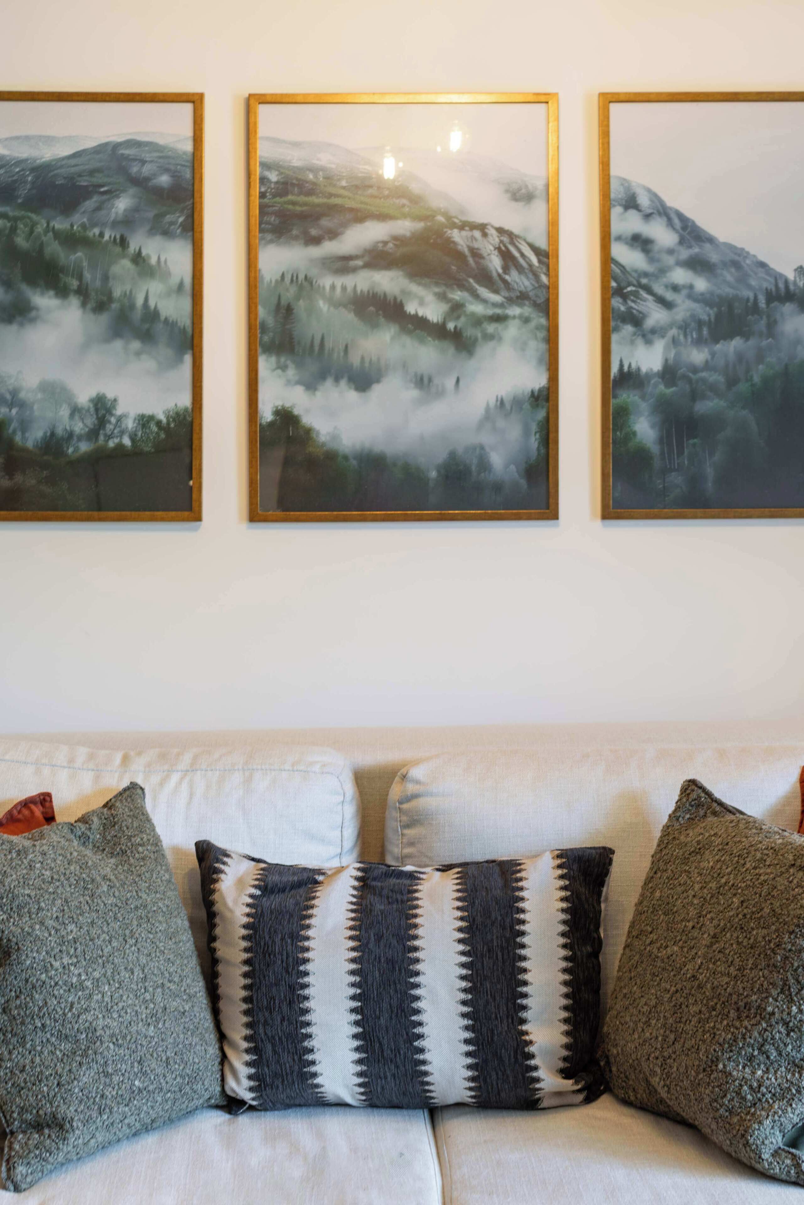 A living room with a white sofa, three framed landscape paintings, and patterned throw pillows.