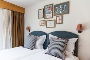 A hotel room with a bed, two pillows, a headboard, a wall with framed pictures, a window with curtains, and a wall-mounted lamp. - Central Verbier | Sleeps 6 | Near Ski Lifts (Val de Bagnes)