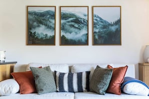 A living room with a white sofa, three framed landscape paintings, and various cushions. - Central Verbier | Sleeps 6 | Near Ski Lifts (Val de Bagnes)
