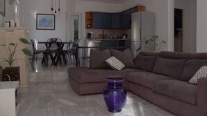 Un salon avec un canapé, une table à manger avec des chaises, un coin cuisine avec des placards et des appareils électroménagers, et un vase violet posé sur le sol.