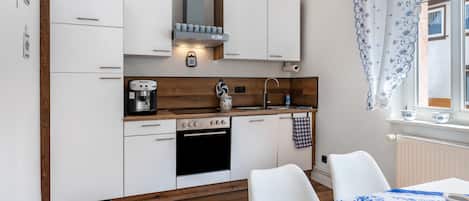 Cocina compacta con armarios blancos, cafetera, lavamanos y ventana con cortinas.