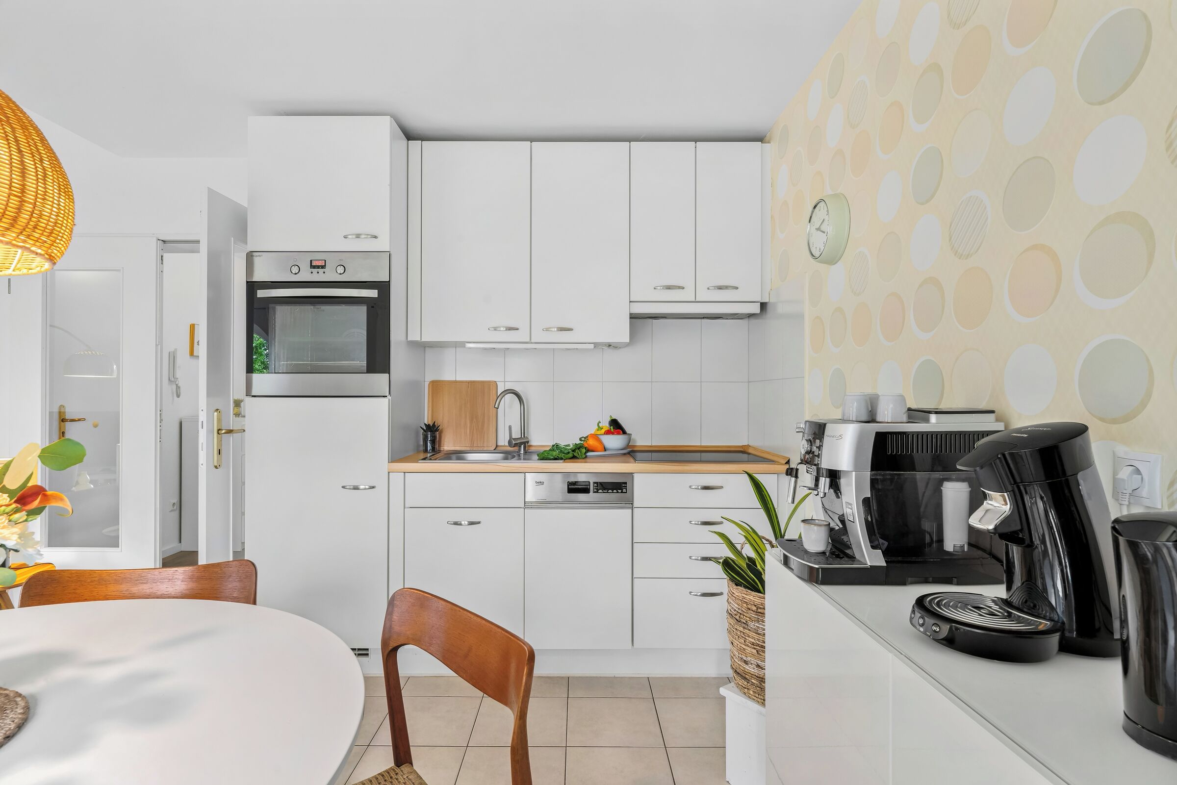 Una cocina moderna con armarios blancos, horno empotrado, cafetera y un comedor con mesa y sillas.