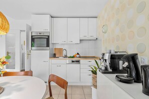A modern kitchen with white cabinets, a built-in oven, a coffee machine, and a dining area with a table and chairs.