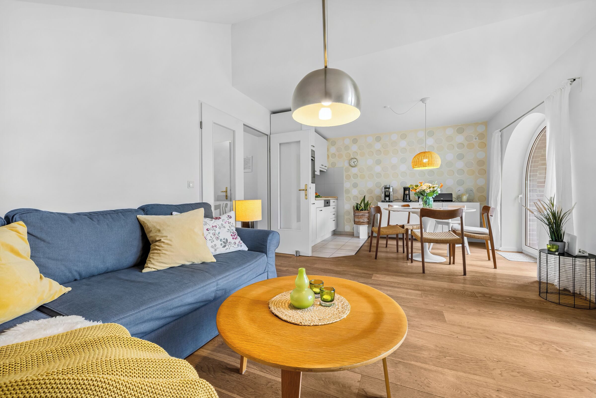 Ein modernes Wohnzimmer mit einem blauen Sofa, einem runden Holztisch und einem Essbereich mit weißem Tisch und Stühlen.