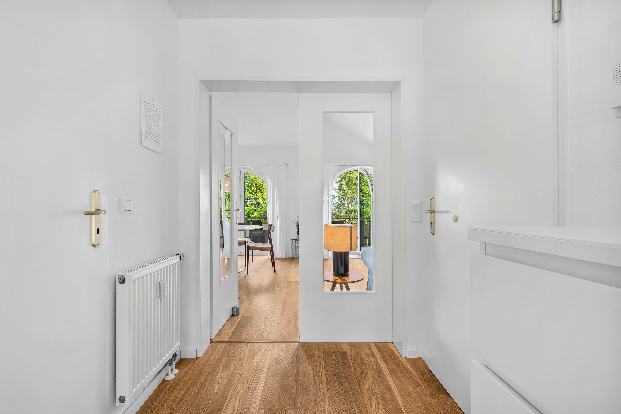 Un pasillo con piso de madera, paredes blancas y una puerta que da a una habitación con vistas al exterior.