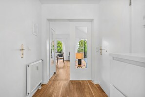 A hallway with a wooden floor, white walls, and a door leading to a room with a view of greenery outside.