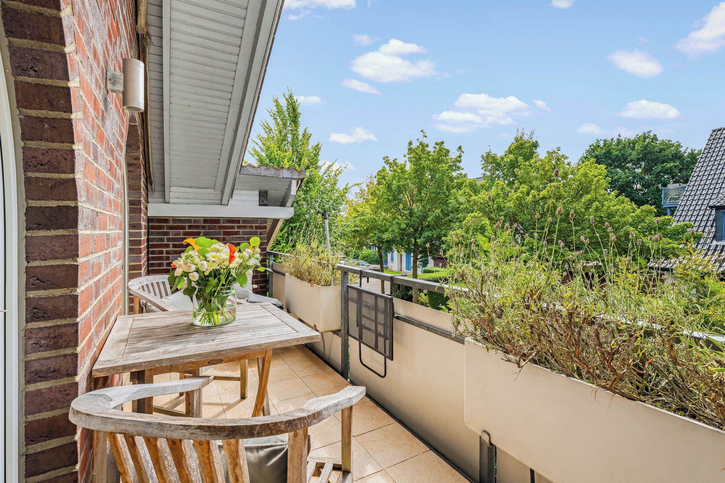 A balcony with a wooden table and chairs, a vase of flowers, and a view of greenery and buildings.