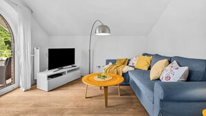 A modern living room with a flat-screen TV on a white stand, a blue sofa with decorative pillows, a wooden coffee table, and a floor lamp. - Nordstern Vacation Rental with Ocean Views and Two Balconies on Schönberg Beach (Schönberg (Holstein))