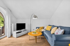 Ein modernes Wohnzimmer mit einem Flachbildfernseher auf einem weißen Schrank, einem blauen Sofa mit Zierkissen, einem Holztisch und einer Stehlampe.