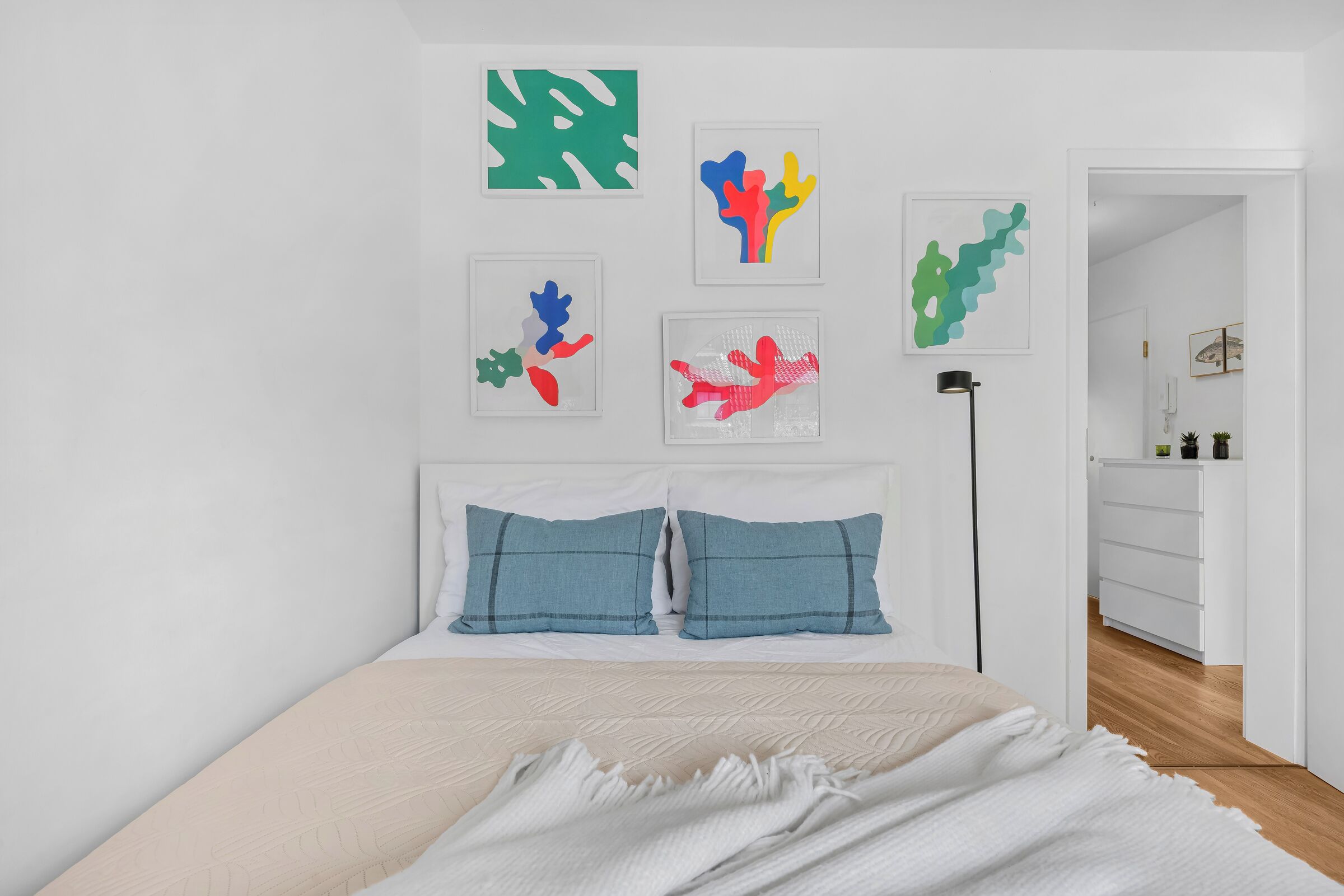 A bedroom with a bed, two pillows, a bedside table, a lamp, and wall art.