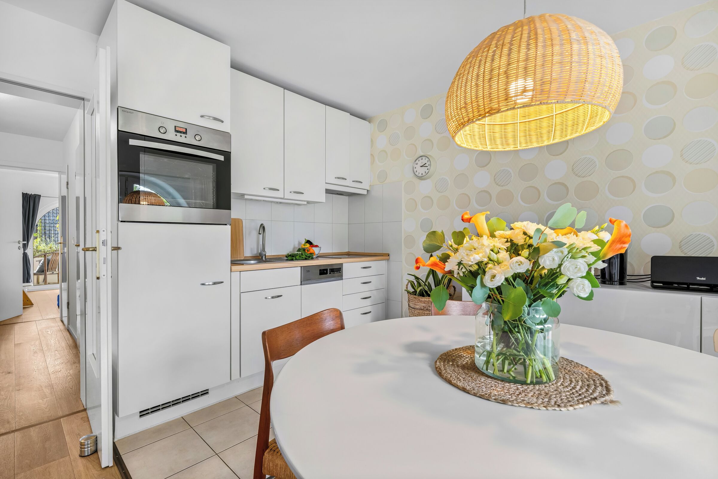 A modern kitchen with white cabinetry, an oven, and a dining area with a round table and a vase of flowers.