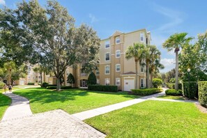 A multi-story residential building with a paved walkway, well-maintained lawn, and mature trees.