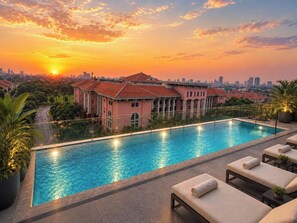 A swimming pool with lounge chairs, a large building with a red roof, and a sunset view.
