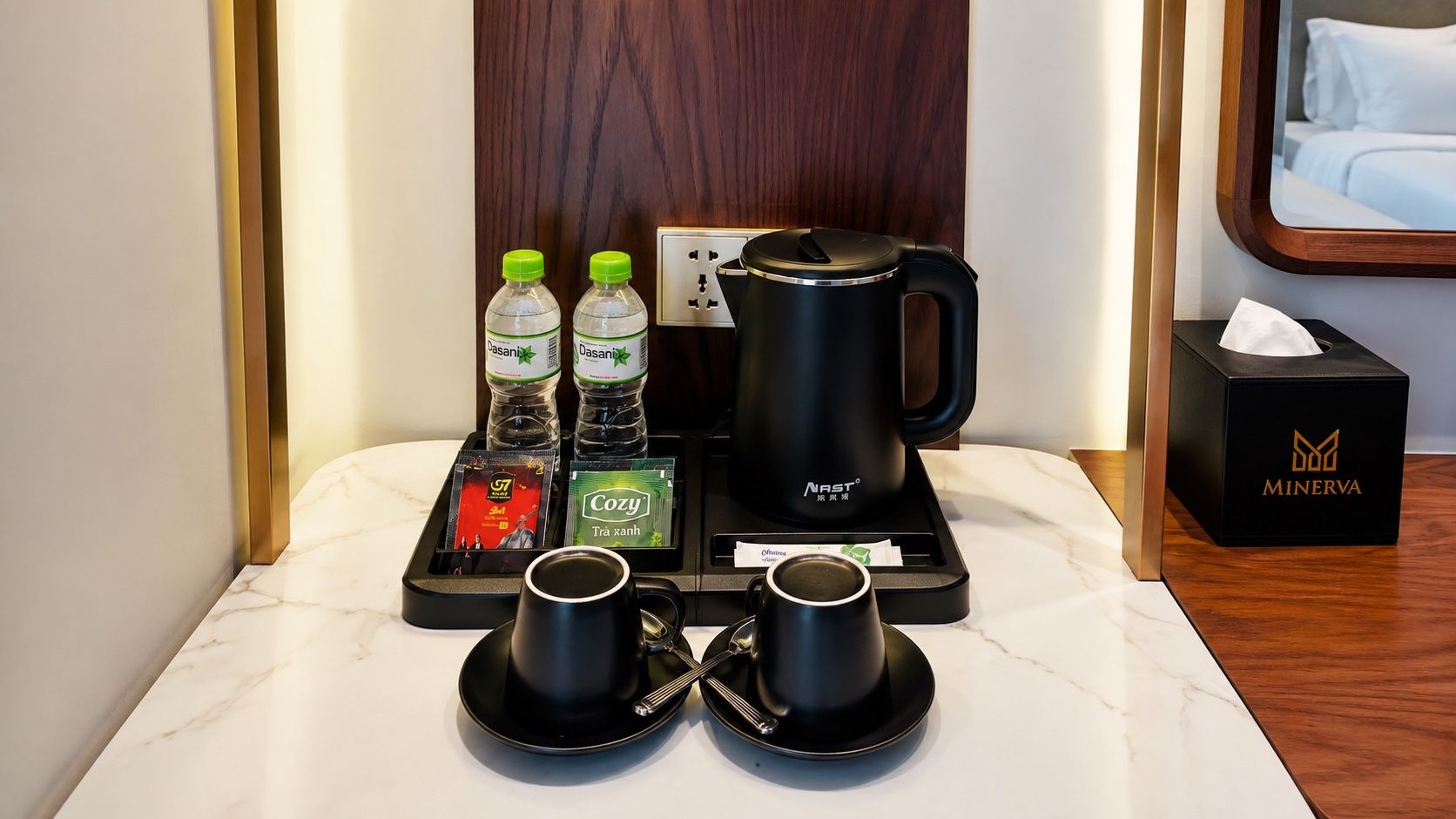 A hotel room with a coffee station including a NAST coffee maker, two Dasani water bottles, a Cozy tea packet, and two black cups on a tray.