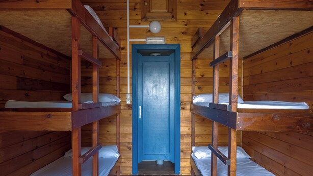 Uma cabana de madeira com beliches dispostos num corredor que leva a uma porta azul.