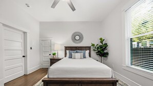 Ein Schlafzimmer mit einem Bett, einem Nachttisch, einer Wanduhr, einem dekorativen Spiegel und einem Fenster mit Jalousien.