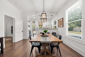 A dining area with a wooden table and chairs, a hanging light fixture, and a view of the outdoors through large windows. - Escape to Gorman Grove—your private Waco sanctuary just 10 mins from Magnolia. <br>Whether you're gathering with family, friends or chasing Chip & Jo dreams, Gorman Grove has historic charm with modern comfort.  Welcome! (Waco)