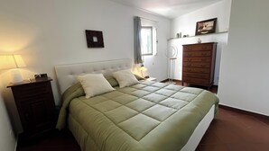 Een slaapkamer met een bed, een dressoir, een nachtkastje, een lamp, een fotolijstje en een raam.
