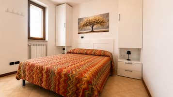 Ein Schlafzimmer mit Bett, Nachttisch, Kommode, Heizkörper, Fenster mit Jalousien und einem an der Wand hängenden Bild von einem Baum.