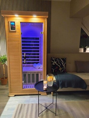 Eine Sauna mit Holzfassade und Glastür, ein kleiner Tisch mit einem Glas Wasser und einem Handtuch, und eine Sitzecke mit einem gemusterten Kissen.