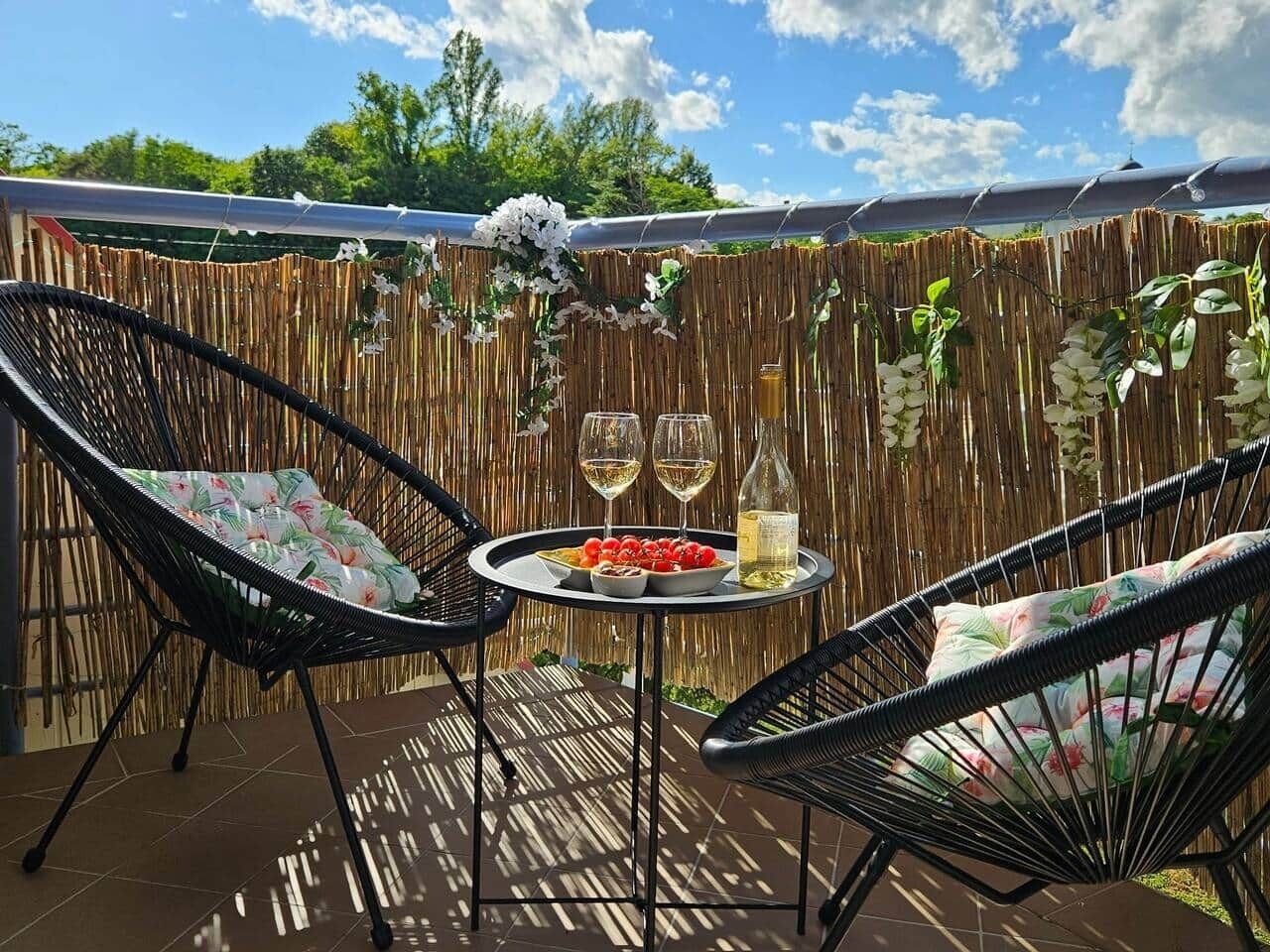 An outdoor patio with two wicker chairs, a round table with a bottle and glasses, and a backdrop of a bamboo fence with hanging plants.