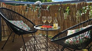Een terras buiten met twee rieten stoelen, een ronde tafel met een fles en glazen, en een achtergrond van een bamboescherm met hangplanten.