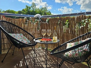 An outdoor patio with two wicker chairs, a round table with a bottle and glasses, and a backdrop of a bamboo fence with hanging plants. - BO - SilvaBlau Apartment Hévíz (Hévíz)