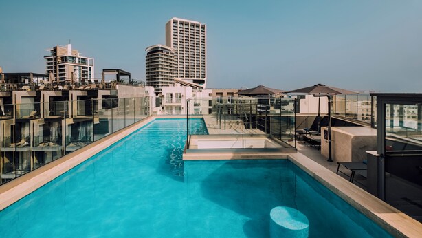 Una piscina en la azotea con barandillas de vidrio, un área de estar y vistas a edificios de la ciudad.